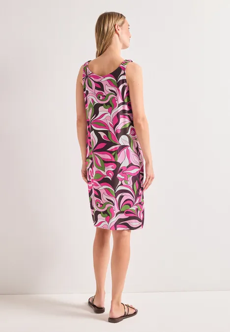 Femme de dos en robe midi sans manches à motif rose, blanc et noir, portant des sandales sur fond clair.