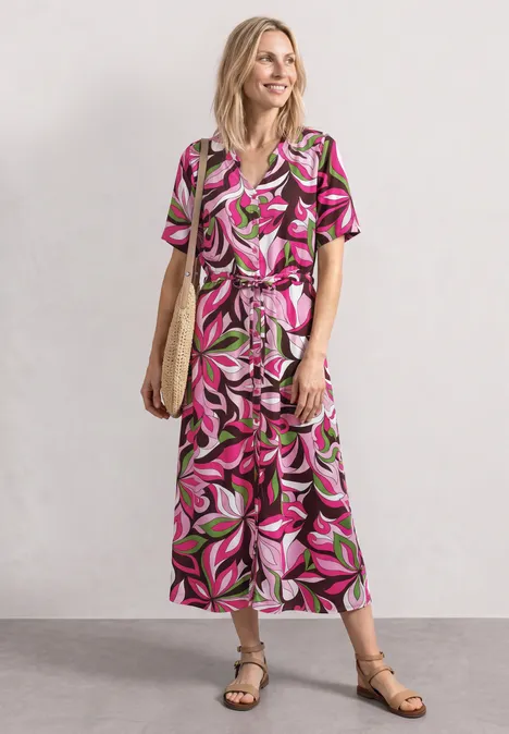 Vrouw in lang, kort mouw kleed met roze-groen bladpatroon, sandalen en strooien tas.