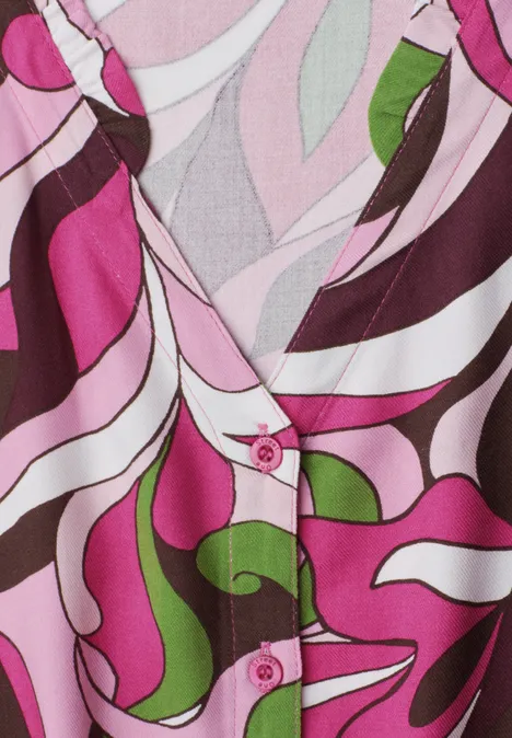 Close-up van een kleurrijk blousepatroon in roze, wit, bruin en groen met V-hals en roze knopen.
