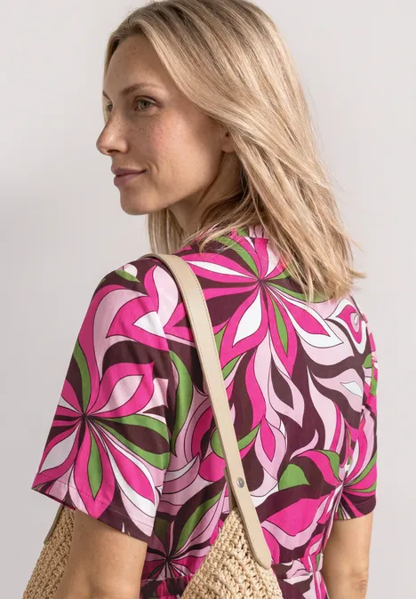 Blonde vrouw draagt een kleurrijk t-shirt met bloemenpatroon en een strooien tas over haar schouder.