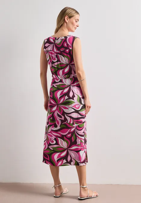 Frau von hinten in einem knielangen, ärmellosen Kleid mit pink-weiß-schwarzem Blattmuster und silbernen Sandalen.