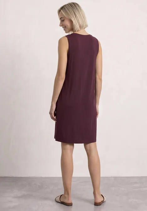 Frau mit kurzen blonden Haaren trägt ärmelloses, knieumspielendes weinrotes Kleid, Ansicht von hinten.
