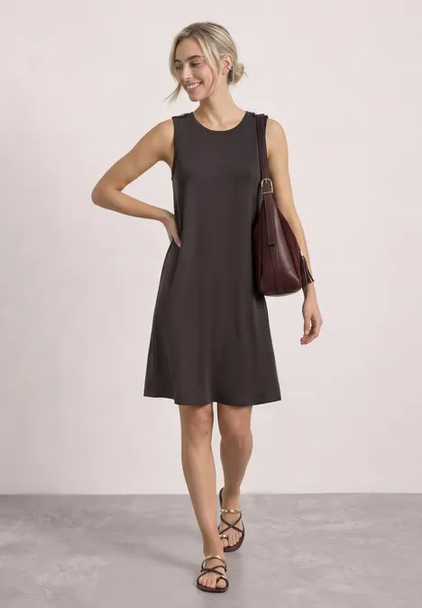 Eine Frau in ärmellosem schwarzem Kleid, Sandalen und Schultertasche steht locker.
