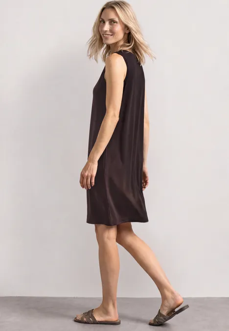Knielanges Kleid mit Rundhals und Knopfdetail black coffee Frau läuft seitlich in ärmellosem schwarzen Kleid und Sandalen vor hellem Hintergrund.
