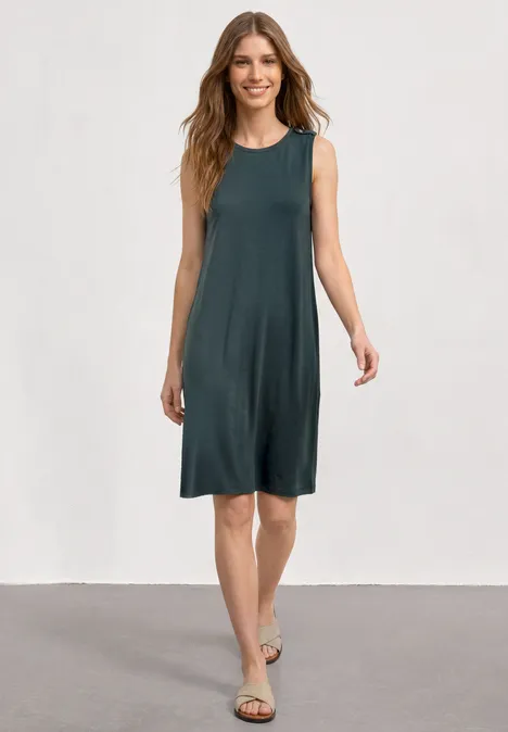 Eine Frau in ärmellosem, knielangem graugrünem Sommerkleid und flachen Sandalen vor neutralem Hintergrund.