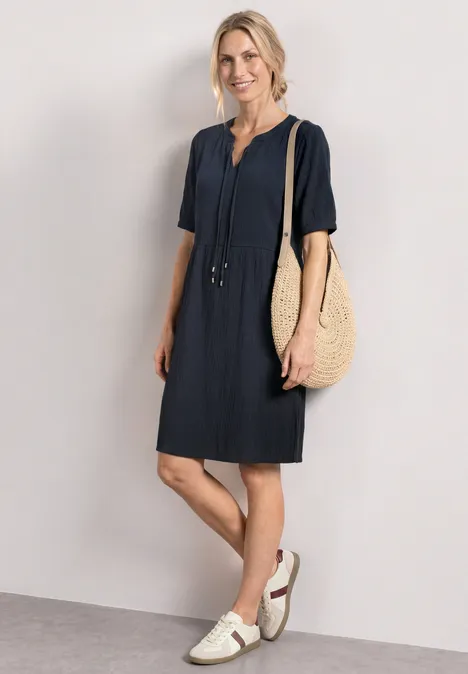 Vrouw in donkerblauwe korte jurk, witte sandalen en met een grote gevlochten tas.