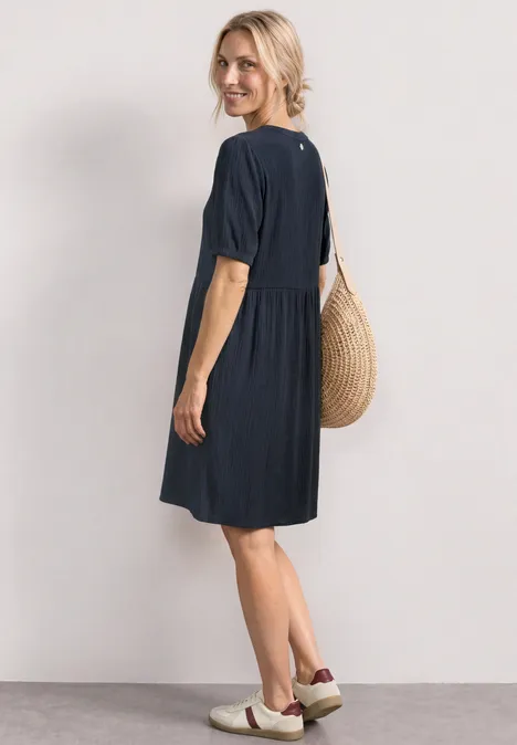 Vrouw in losse marineblauwe jurk met korte mouwen, gevlochten tas en slip-on schoenen, zijwaarts.