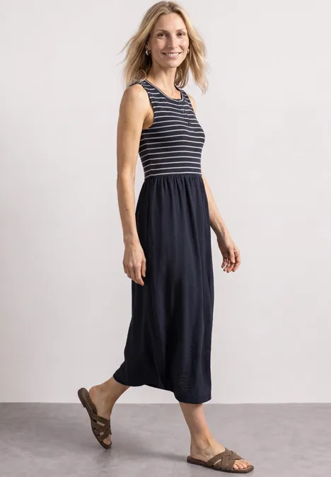 Une femme souriante porte un débardeur rayé bleu marine et une jupe mi-longue avec des sandales.