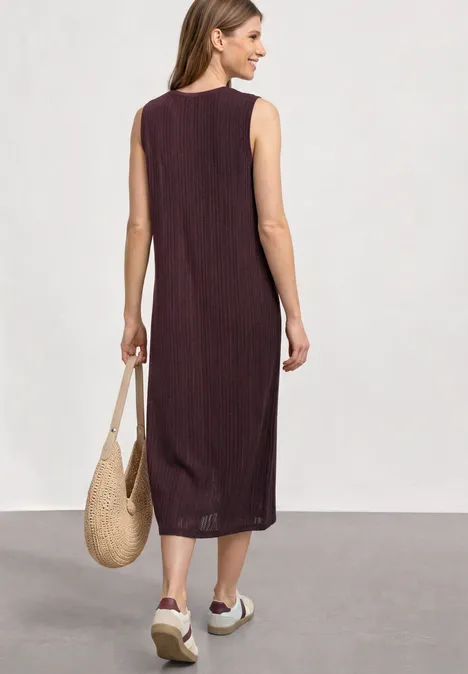 Frau von hinten in ärmellosem, midilangen, dunkelrotem Kleid, mit Strohtasche und weißen Plateauschuhen.