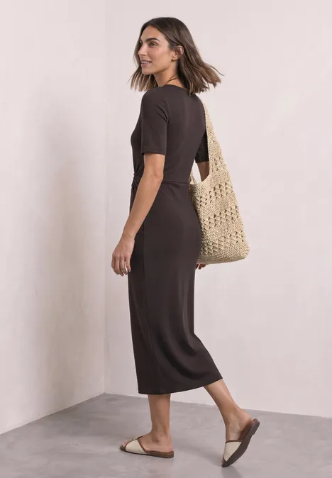 Une femme en robe midi marron, sac en paille tissé à l'épaule et sandales plates.