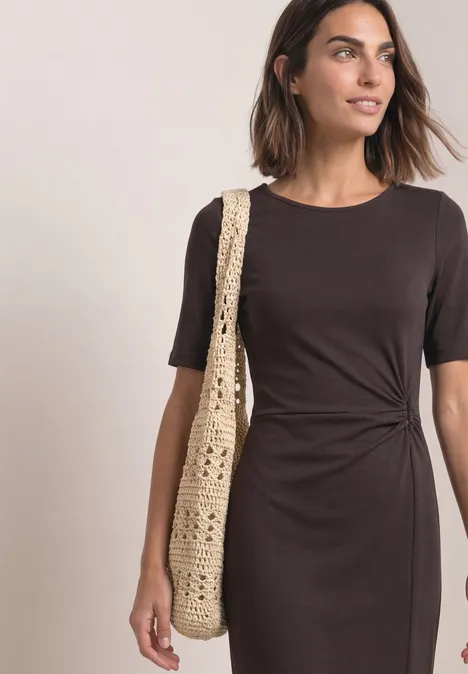 Femme en robe marron mi-longue avec noeud latéral et manches courtes, portant un sac en crochet.