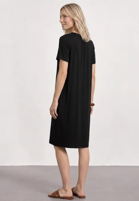 Vrouw met blond haar in een zwart midi-jurkje en sandalen, van achteren voor een witte muur.