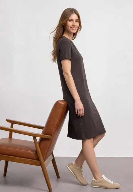 Vrouw in donkergrijze, knielange jurk en sneakers, schuin leunend tegen een houten leren fauteuil voor een witte muur.
