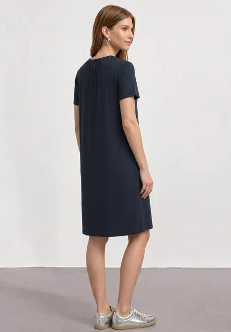 Vrouw van achteren in een donkerblauwe knielange jurk en zilverkleurige instappers, neutrale achtergrond.