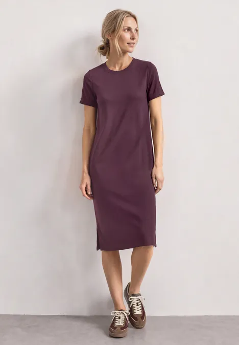 Frau trägt schlichtes violettes T-Shirt-Kleid bis zur Wade und Sneaker vor neutralem Hintergrund.