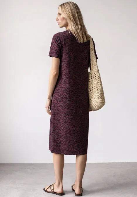 Midi-jurk met grafisch patroon rustic rouge Vrouw in midi-jurk met patroon, draagt rieten tas en platte sandalen, staat zijwaarts voor lichte achtergrond.