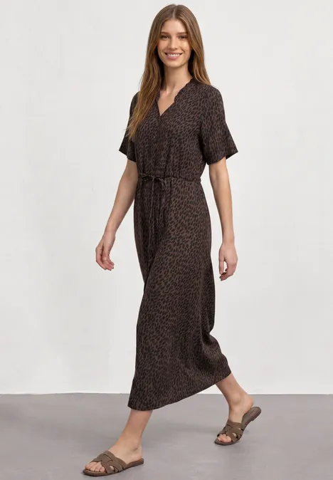 Vrouw in een bruine maxi-jurk met ceintuur en sandalen, die glimlachend loopt.