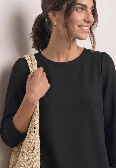 Knielanges Kleid mit Rundhals Black Frau lächelt in schwarzem Oberteil und trägt eine beige gehäkelte Tasche über der Schulter.