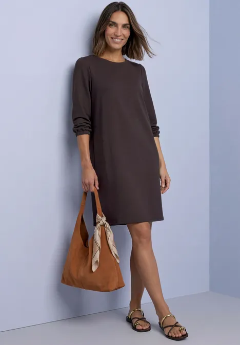 Knielanges Kleid mit Rundhals ruby vine Frau in lockerem, braunem Kleid mit lässiger Handtasche und Sandalen vor pastellfarbener Wand.