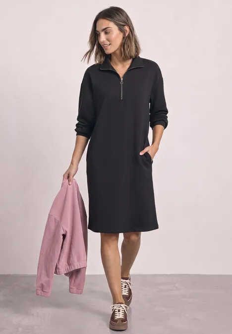 Frau im schwarzen knielangen Kapuzenkleid mit Reißverschluss hält rosa Jacke in der Hand.