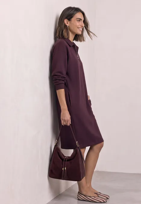 Robe en molleton arrivant aux genoux, avec fermeture éclair rustic rouge Femme de profil portant une robe-chemise violet foncé, un sac à épaule assorti et des sandales rayées.