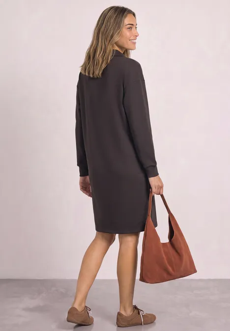 Robe en molleton arrivant aux genoux, avec fermeture éclair black coffee Femme vue de dos portant une robe sombre mi-longue, sac cabas marron et mocassins beiges.