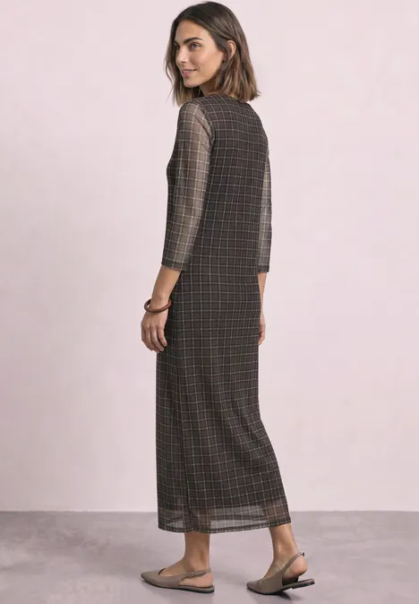 Frau in langem kariertem Maxikleid mit 3/4-Ärmeln und flachen Sandalen, seitlich stehend.