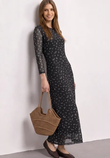 Eine Frau trägt ein langes schwarzes Kleid mit floralem Muster und hält eine Korbtasche.