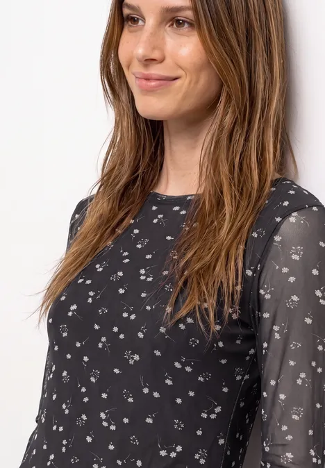 Frau in schwarzem, leicht transparentem Oberteil mit kleinem weißen Blumenmuster und dezentem Lächeln.