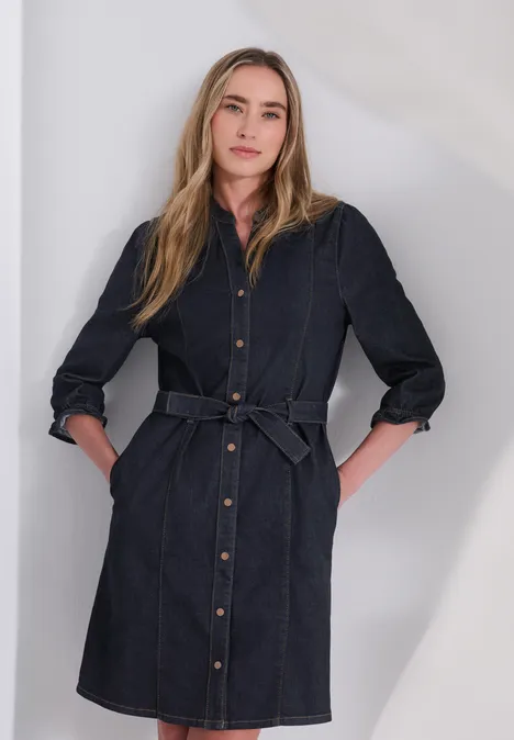 Femme en robe-chemise en jean bleu, ceinture à la taille, mains dans les poches, fond neutre.