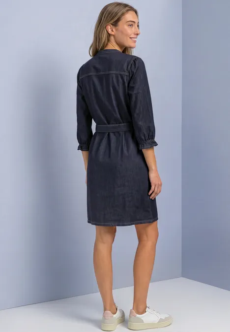 Femme vue de dos portant une robe chemise en jean bleu foncé avec manches 3/4, ceinture et baskets claires.