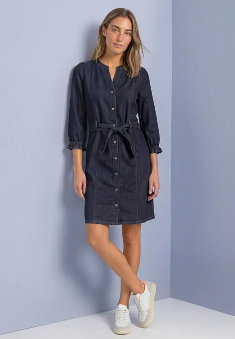 Une femme porte une robe-chemise en jean foncé ceinturée et des baskets blanches, debout devant un mur clair.