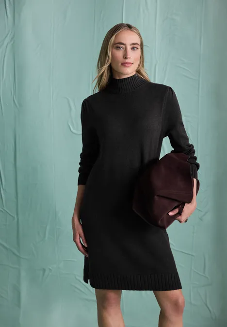 Frau in schwarzem Strickkleid und Rollkragen, hält eine bordeauxrote Tasche vor grünem Hintergrund.
