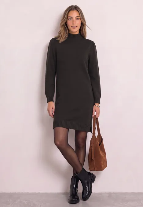 Frau trägt dunkles, langärmeliges Kleid, schwarze Strumpfhose, Boots und braune Schultertasche vor hellem Hintergrund.