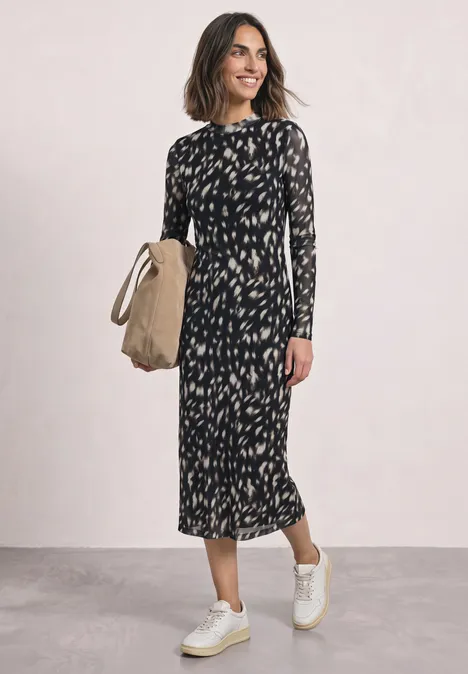 Frau trägt ein schwarzes, gemustertes Midikleid mit langen Ärmeln, weiße Sneaker und hält eine Strohtasche.