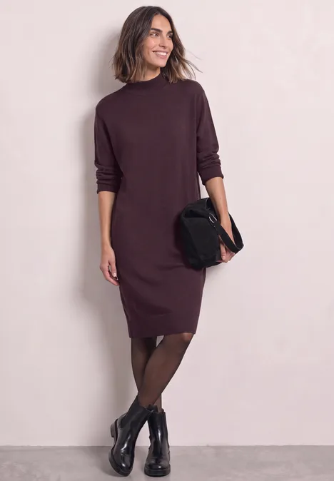Model in bordeauxrotem Strickkleid mit langen Ärmeln, schwarzen Strumpfhosen, Stiefeletten und Clutch.