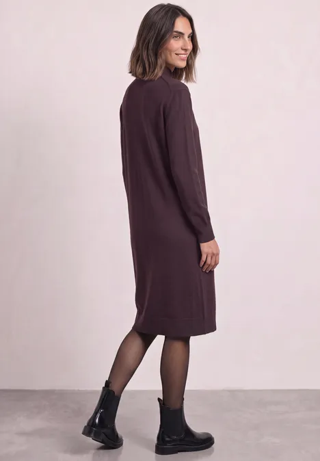 Frau in knielangem, langärmeligen, dunkelviolettem Kleid mit schwarzen Boots und Strumpfhose, seitlich stehend.