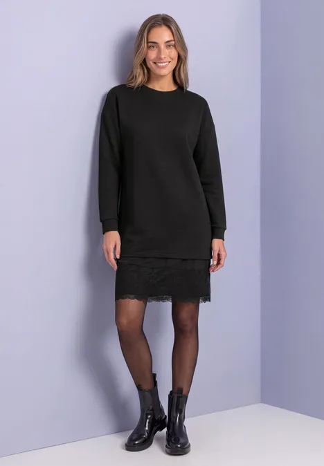 Knielanges Sweatkleid mit Spitzenkante Black Frau in schwarzem Strickkleid mit Spitzenbesatz, schwarzen Strumpfhosen und Stiefeletten vor hellvioletter Wand.