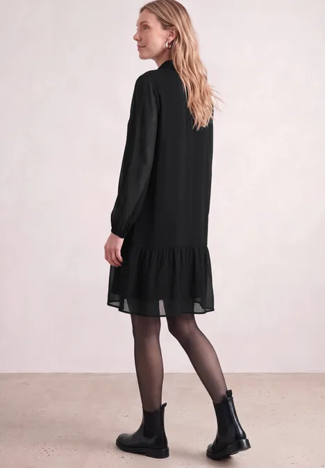 Frau in seitlicher Ansicht trägt schwarzes, lockeres Kleid mit Volant am Saum, schwarze Stiefeletten und Strumpfhose.