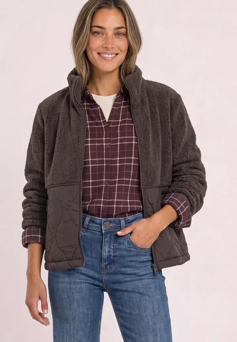 Lächelnde Frau trägt braune Fleecejacke über kariertem Hemd und blaue Jeans.