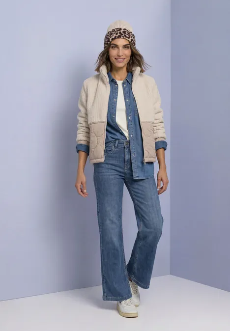 Frau in Jeansoutfit mit heller Jacke und gemustertem Hut vor violettem Hintergrund.