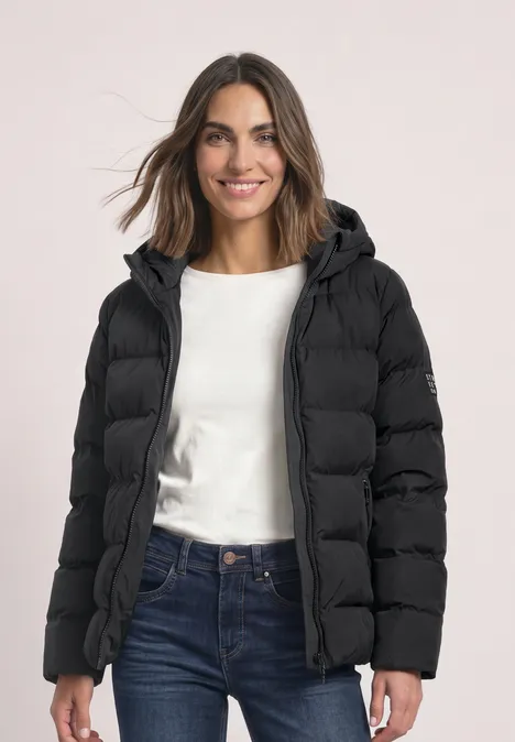 Vrouw met zwarte pufferjas met capuchon, wit shirt en blauwe jeans lacht.