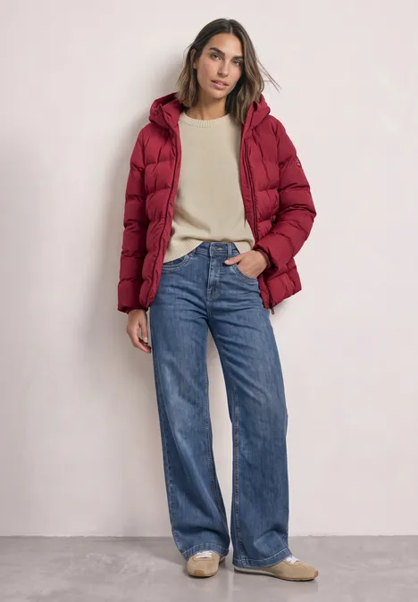Kort jasje met gelijmde stiksels vintage red Vrouw in rode gewatteerde jas, beige trui en wijde jeans draagt lichtbeige schoenen.