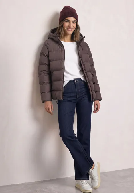 Femme portant une doudoune marron, jean évasé, t-shirt blanc et bonnet, posant décontractée contre un mur.