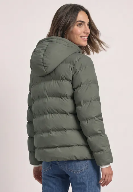 Frau in khakigrüner Steppjacke mit Kapuze und Jeans, schaut lächelnd über die Schulter.