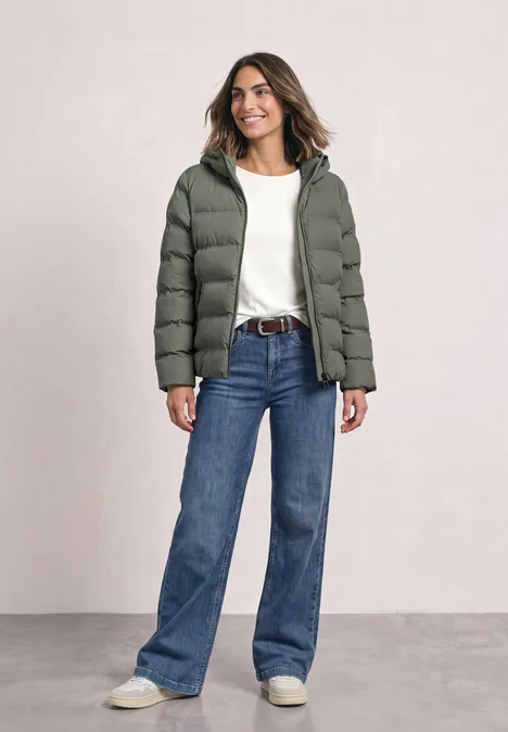Frau in grüner Steppjacke, weißem Shirt und blauen Jeans steht lächelnd vor neutralem Hintergrund.