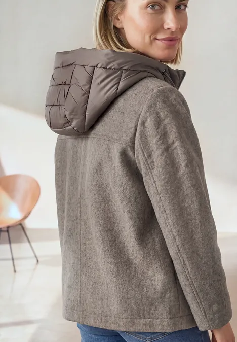 Frau trägt graubraune Jacke mit Kapuze, schaut über die Schulter in minimalistischer Wohnumgebung.