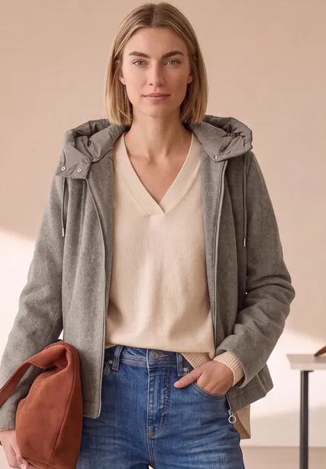 Frau mit grauer Jacke und cremefarbenem Pullover vor neutralem Hintergrund