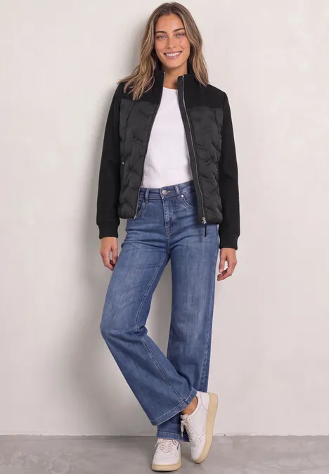 Frau in schwarzer Steppjacke, weißem T-Shirt, blauen Jeans und weißen Sneakern, lächelnd vor hellem Hintergrund.