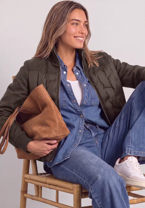 Jeune femme souriante en chemise et jean, veste matelassée verte, baskets blanches, assise sur un tabouret, sac marron.